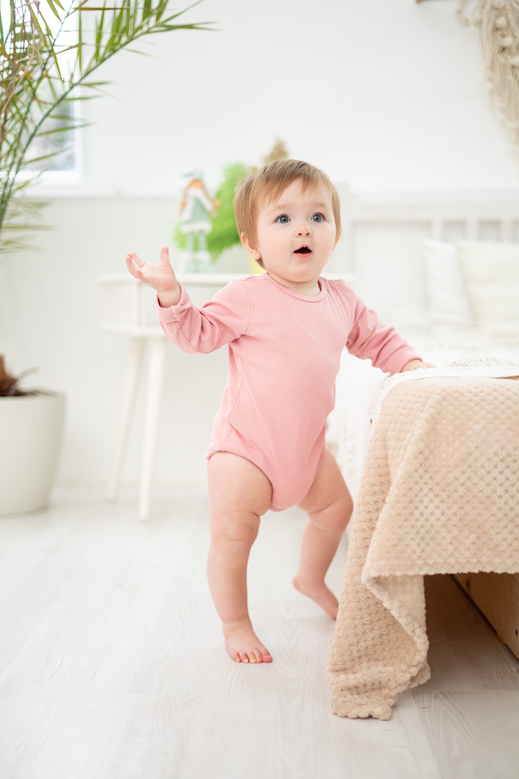 a cute healthy girl is standing by the bed in the bedroom at home, the baby's first steps, the baby is learning to walk Montessori Kinder