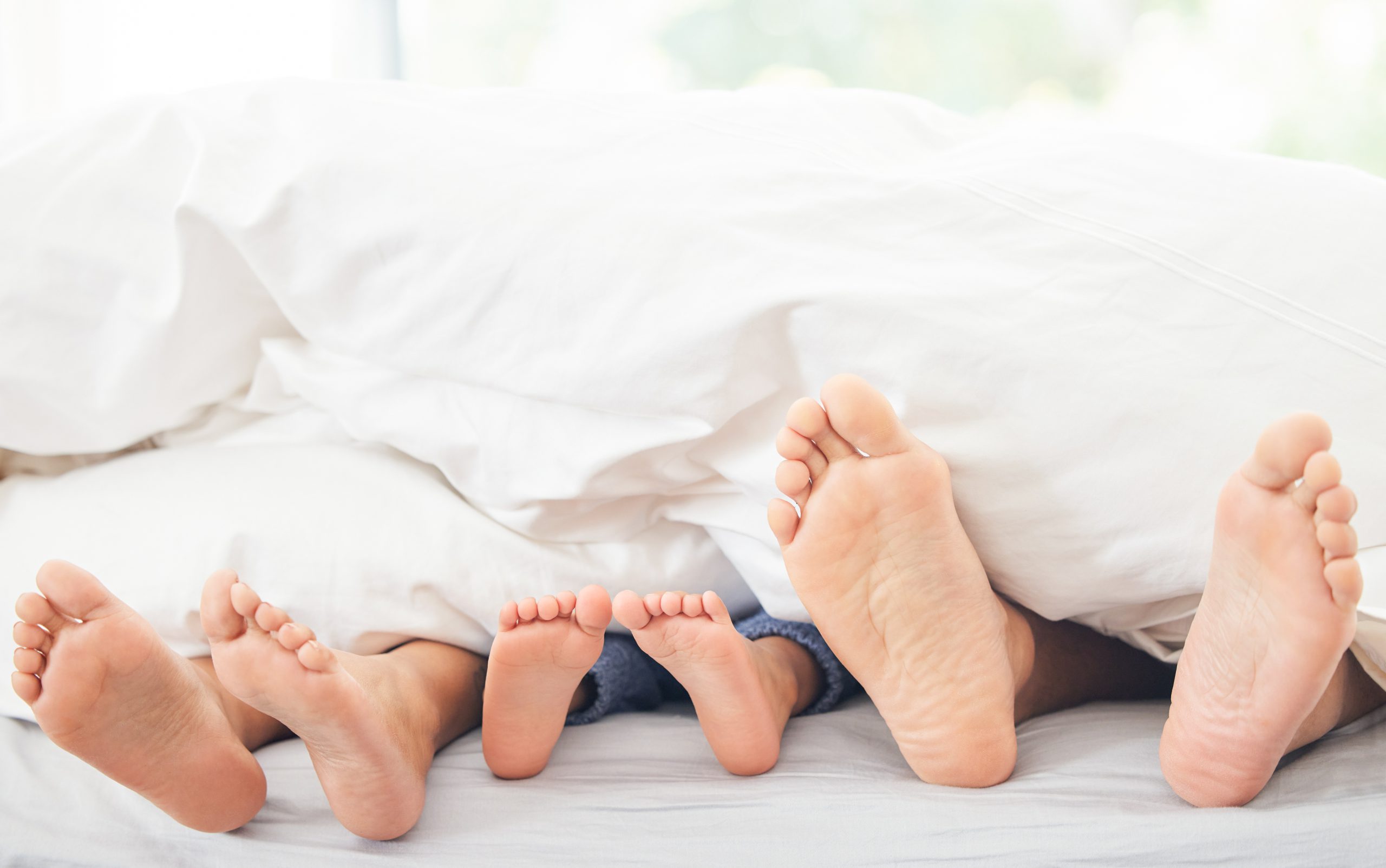 Montessori im Alltag: Kinderfüße und Elternfüße unter weißer Bettdecke – gemeinsame Morgenzeit, Nähe und Ruhe im Familienalltag.